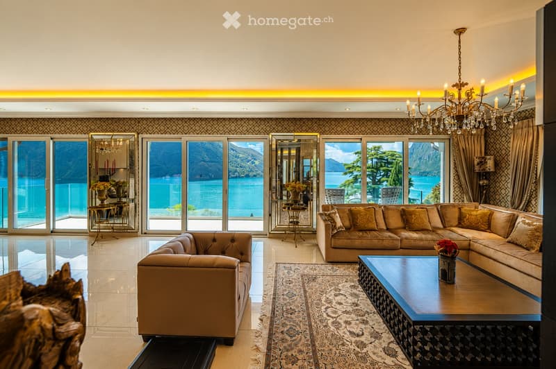 Elegant living room with large windows offering a scenic lake view, featuring a beige sectional sofa, coffee table, chandelier, and decorative mirrors.