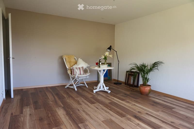 Furnished Room. A cozy, well-lit room with wooden flooring, a white wicker chair with cushions, a small white table holding a vase with flowers, a bottle, and a glass, a floor lamp, and a potted plant near the wall.