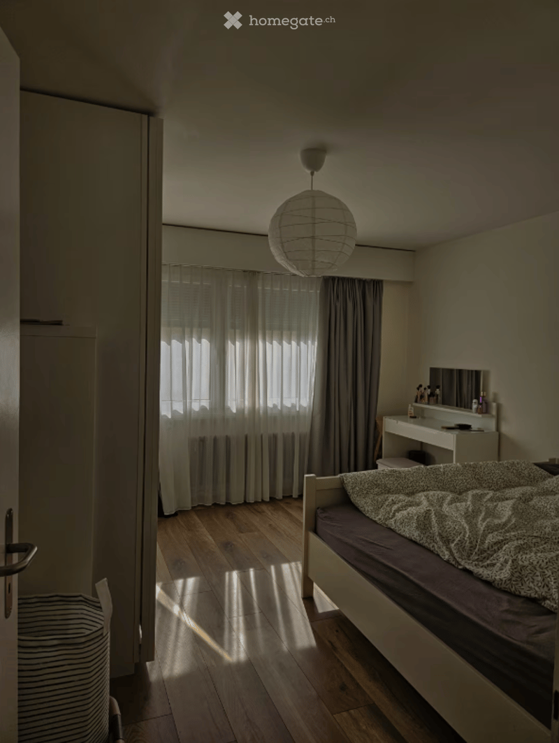 Bedroom. A modern, minimalist bedroom with a wooden floor, a single bed with a patterned duvet, a vanity with a mirror, and a hanging spherical lamp. The room features large windows with sheer curtains allowing natural light to filter in.