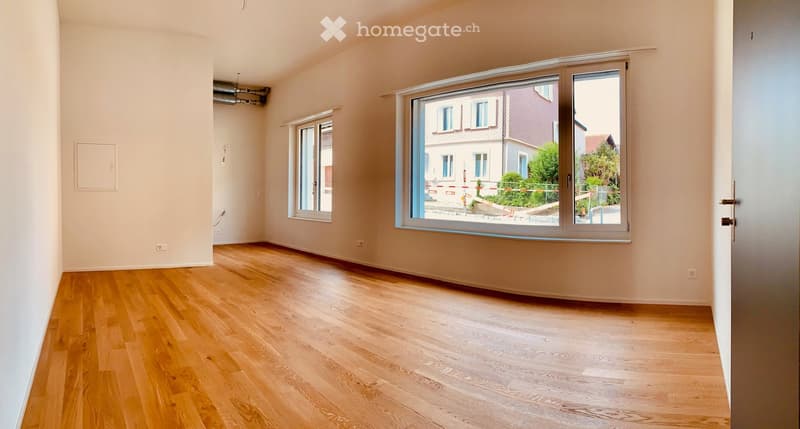 Empty room with light wooden flooring, white walls, and large windows offering a view of the street and neighboring buildings.