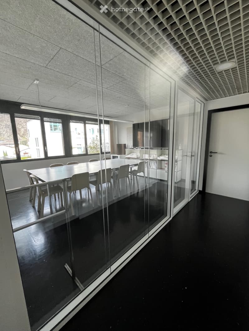 Modern office space with a large conference table, chairs, a screen, and a shelf with books and office supplies. The room features large windows and a glass partition.