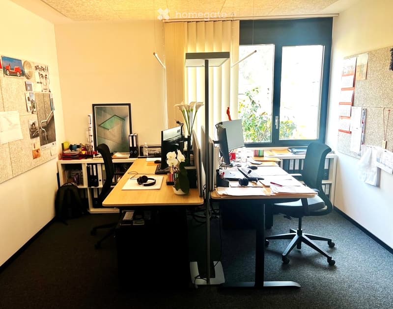 Office space with two desks, chairs, computers, screens, keyboards, mice, headphones, a flower vase, calendar, picture frame, and a backpack.