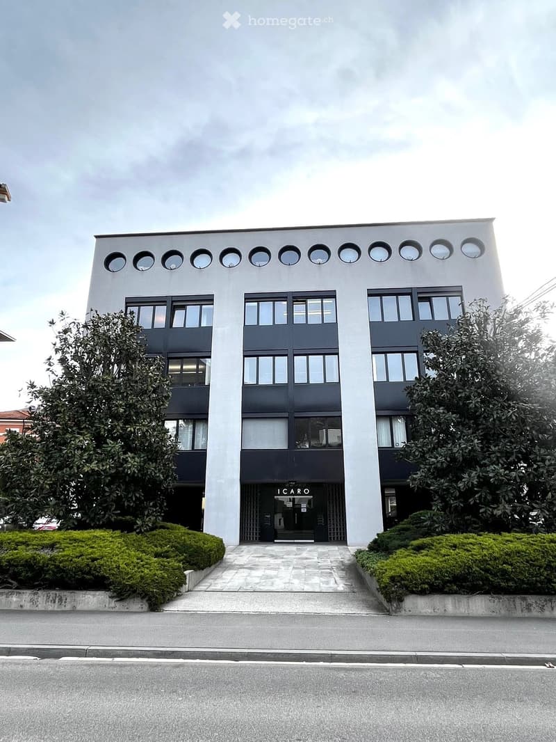 Modern two-story building with white facade, circular windows on the roof, and large rectangular windows on the lower level. The entrance features a black sign with the text "ICARO" and is flanked by greenery and a paved walkway.