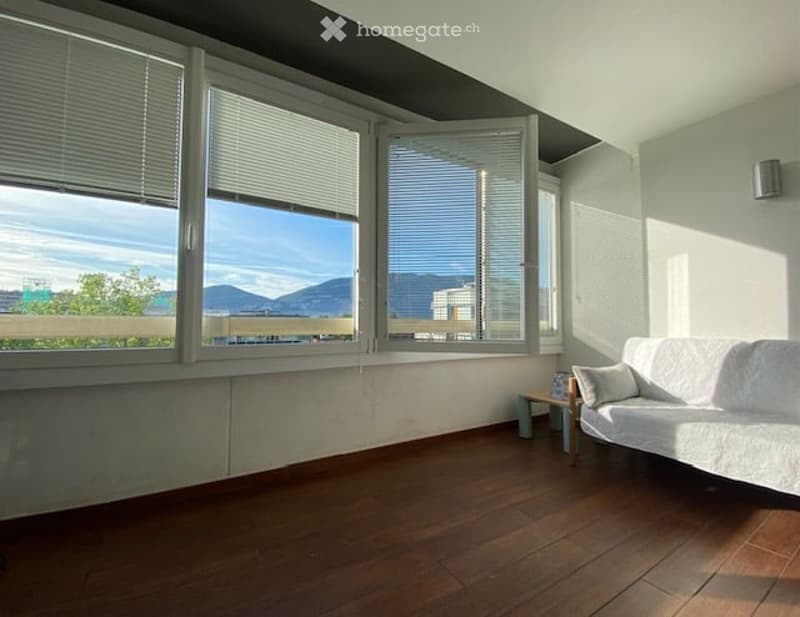 Bright living room with large windows offering a scenic view, featuring a white sofa, small wooden table, and chair.