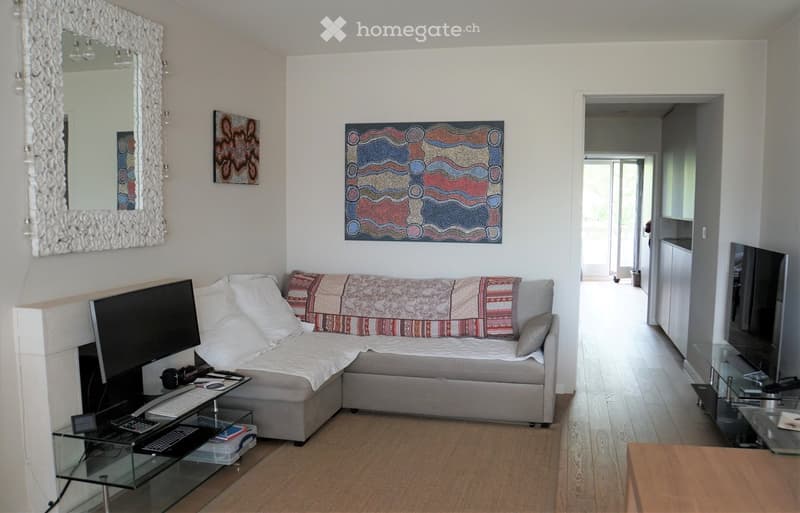 Modern living room with a grey sectional sofa, glass coffee table, laptop, keyboard, artwork, and a large mirror on the wall.