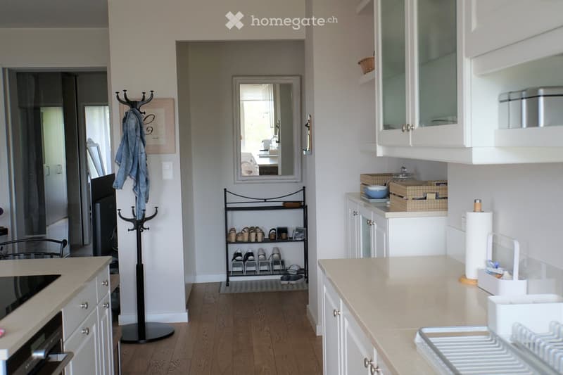 A modern, well-lit hallway and kitchen area with white cabinets, a light-colored countertop, a shoe rack, a coat rack, and a mirror. The floor is wooden, and the space is tidy and organized.