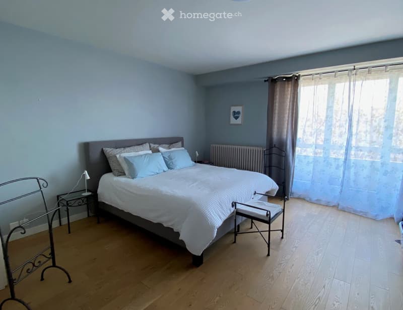 Cozy bedroom with a large bed, nightstand, lamp, and a small chair near the window. Light blue walls and wooden flooring.