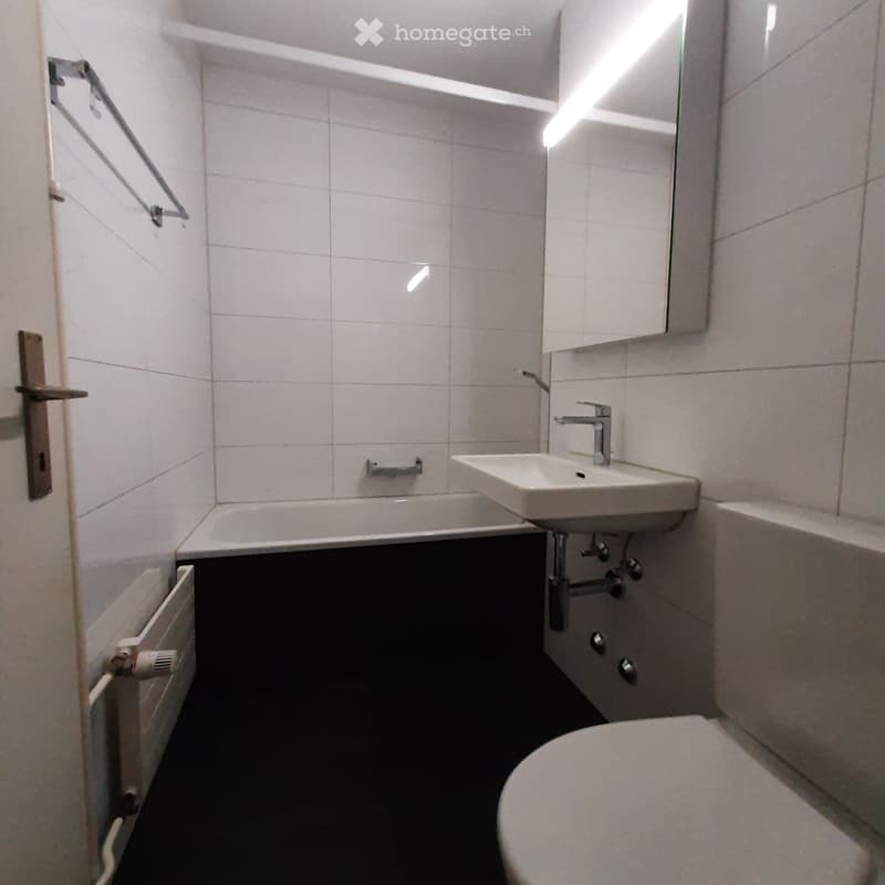 Modern bathroom with white tiles, a sink with a modern faucet, a bathtub, a toilet, and a mirror above the sink. A radiator is visible on the left wall.