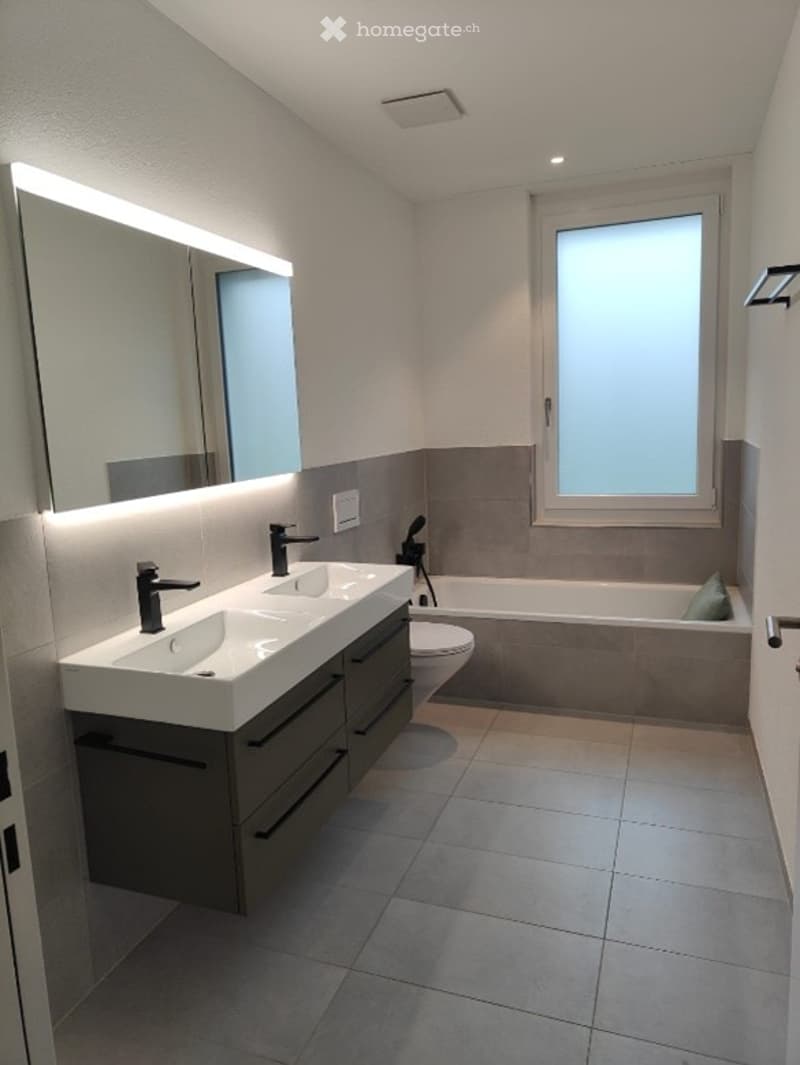 Modern bathroom with double sink, large mirror, bathtub, and toilet. Features tiled walls and floor, and a window providing natural light.