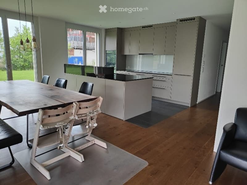Open-plan kitchen and dining area with modern grey cabinets, a large wooden dining table surrounded by chairs, and pendant lights hanging from the ceiling. The space features hardwood flooring and large windows offering a view of the outdoors.
