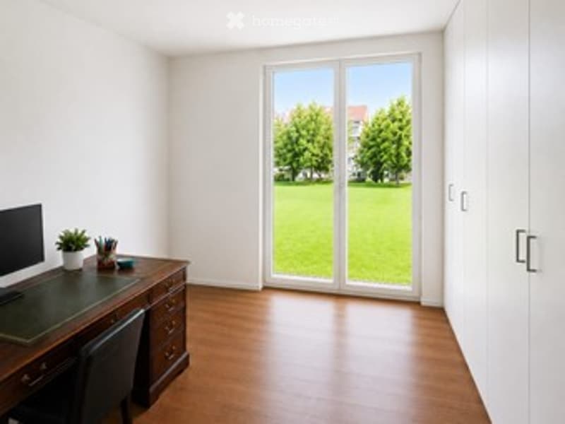 Bright, unfurnished room with wooden flooring, large window with a view of a green lawn, and a small desk with a computer and plant.