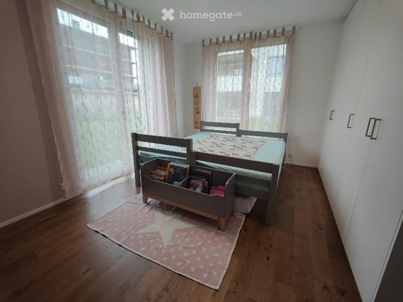 Cozy bedroom with wooden flooring, a single bed with a colorful quilt, a built-in bookshelf under the bed holding books, and a pink star-patterned rug.