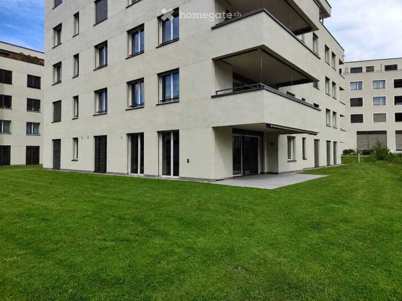 Modern multi-story residential building with balconies and large windows, surrounded by a well-maintained lawn.