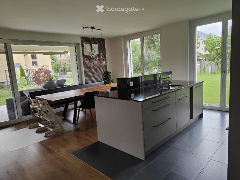 Open-plan living and dining area with a modern kitchen island, large windows offering a view of the garden, and contemporary furniture including a sofa, dining table, and chairs.