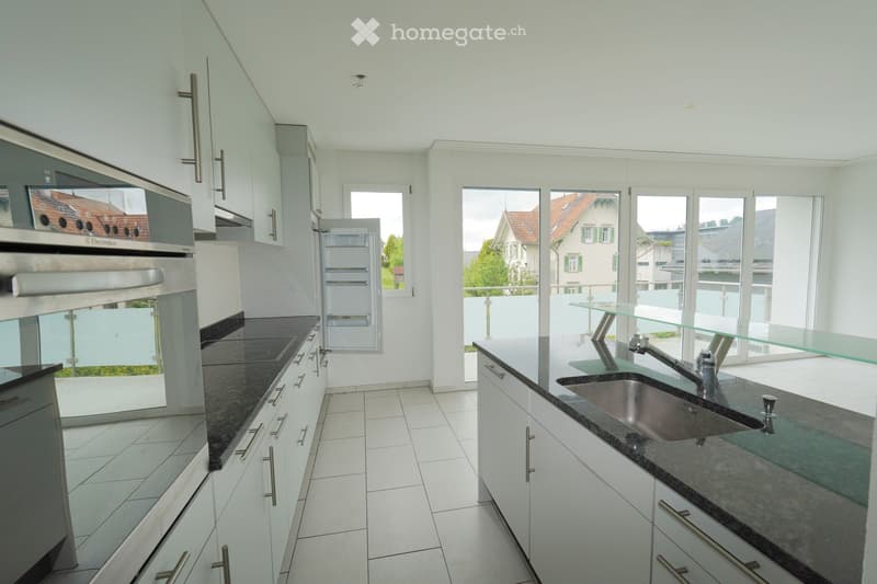 Modern kitchen with white cabinets, stainless steel appliances, dark countertops, and large windows overlooking a balcony.