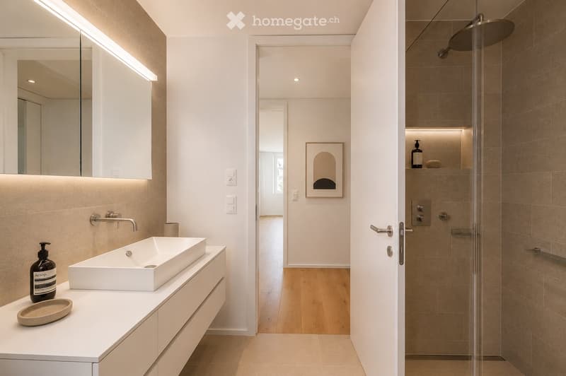 Modern bathroom with a white sink, mirror, and shower area. Features a sleek design with built-in lighting and a glass-enclosed shower.