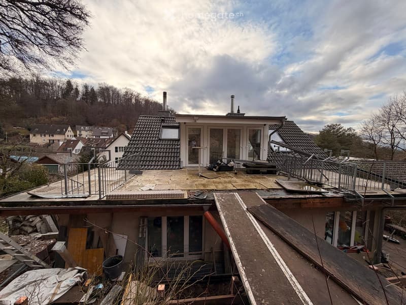 Roof of a house with exposed tiles, metal railings, and construction materials scattered around. The surrounding area includes neighboring houses and a hilly landscape.