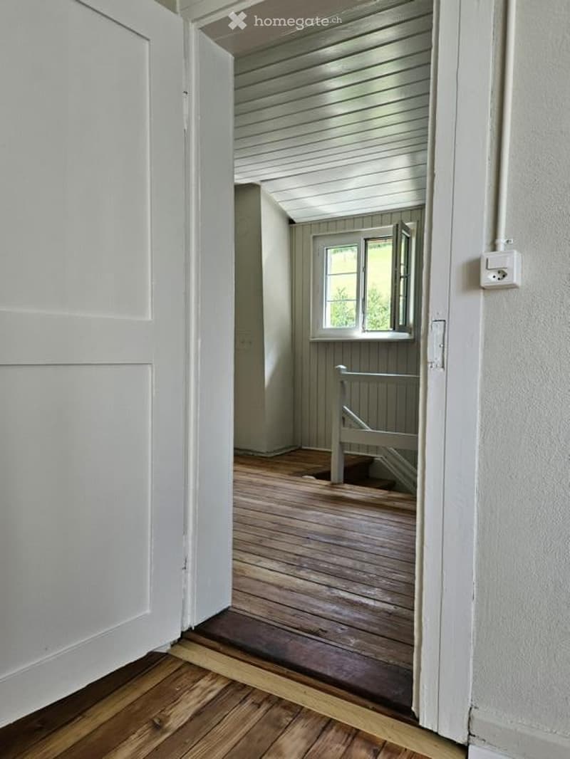 A narrow hallway with wooden flooring and white walls, leading to a small room with a window and a simple wooden bench.