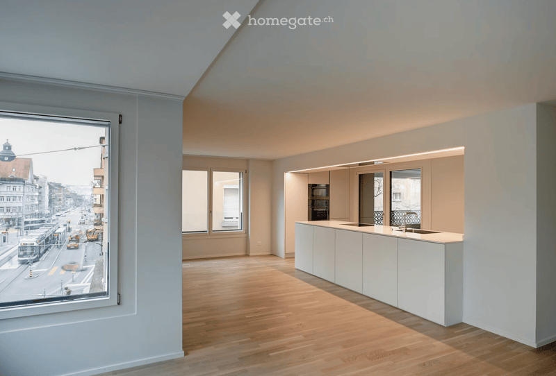 Modern, minimalist kitchen with a white island, sink, and built-in cabinets. The space features light wood flooring and large windows offering a city view.