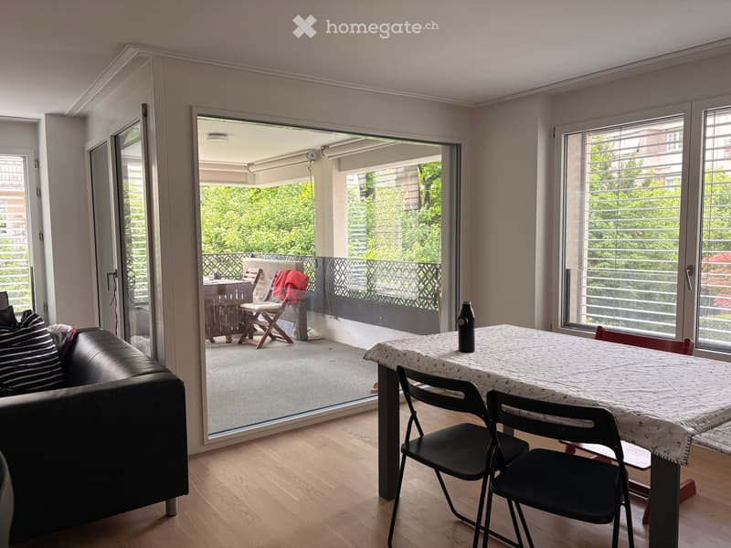 Modern living room with wooden flooring, large sliding glass doors leading to a balcony, sofa, dining table, and chairs.