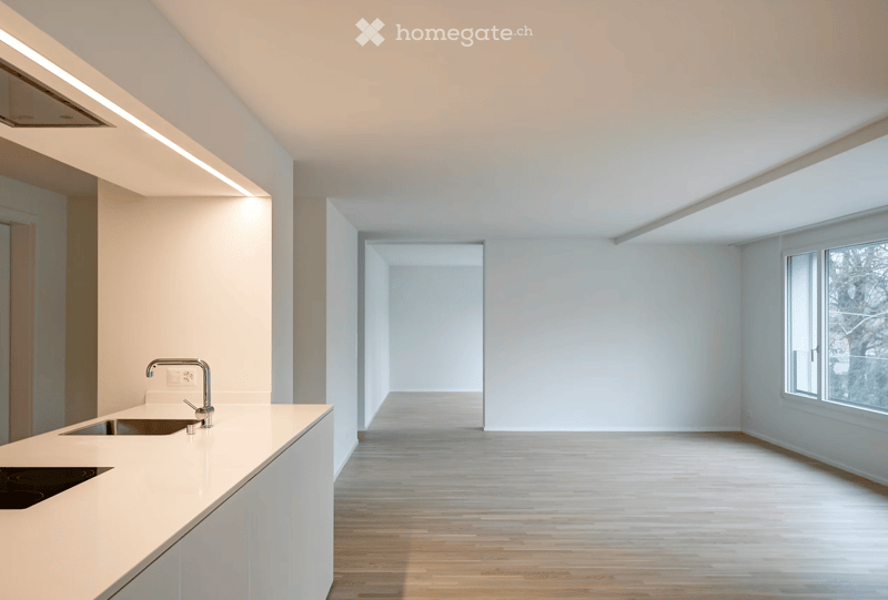 Modern, minimalist kitchen with white countertops, a built-in sink, and a sleek faucet. The space features light wooden flooring, large windows, and recessed lighting.