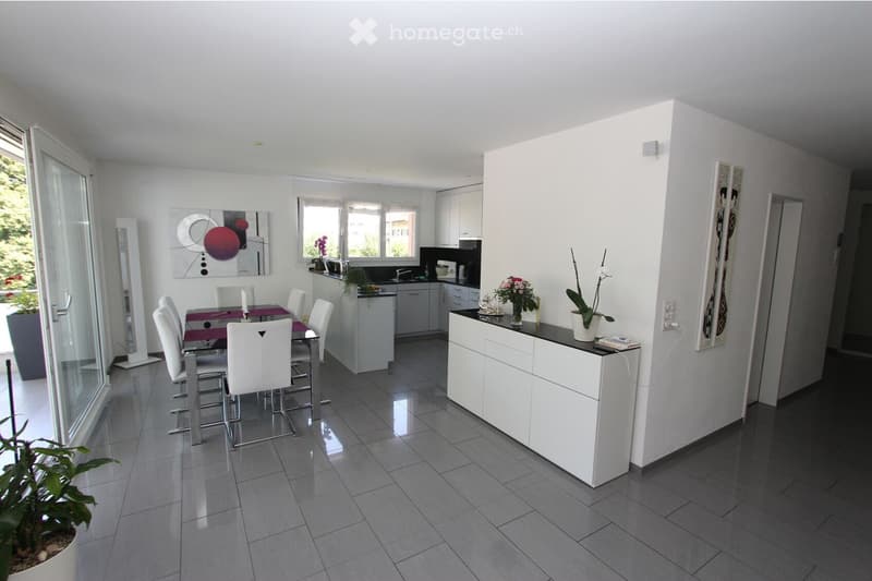 Modern, open-plan dining and kitchen area with white cabinets, a dining table with chairs, and a kitchen counter with a vase of flowers.