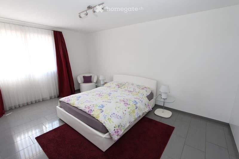 Modern bedroom with a white bed, floral duvet, red throw rug, white side chair, and two table lamps.