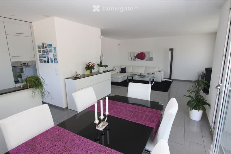 Modern open-plan living and dining area with white walls, grey tiled flooring, white sofa, glass coffee table, black dining table with white chairs, and potted plants.