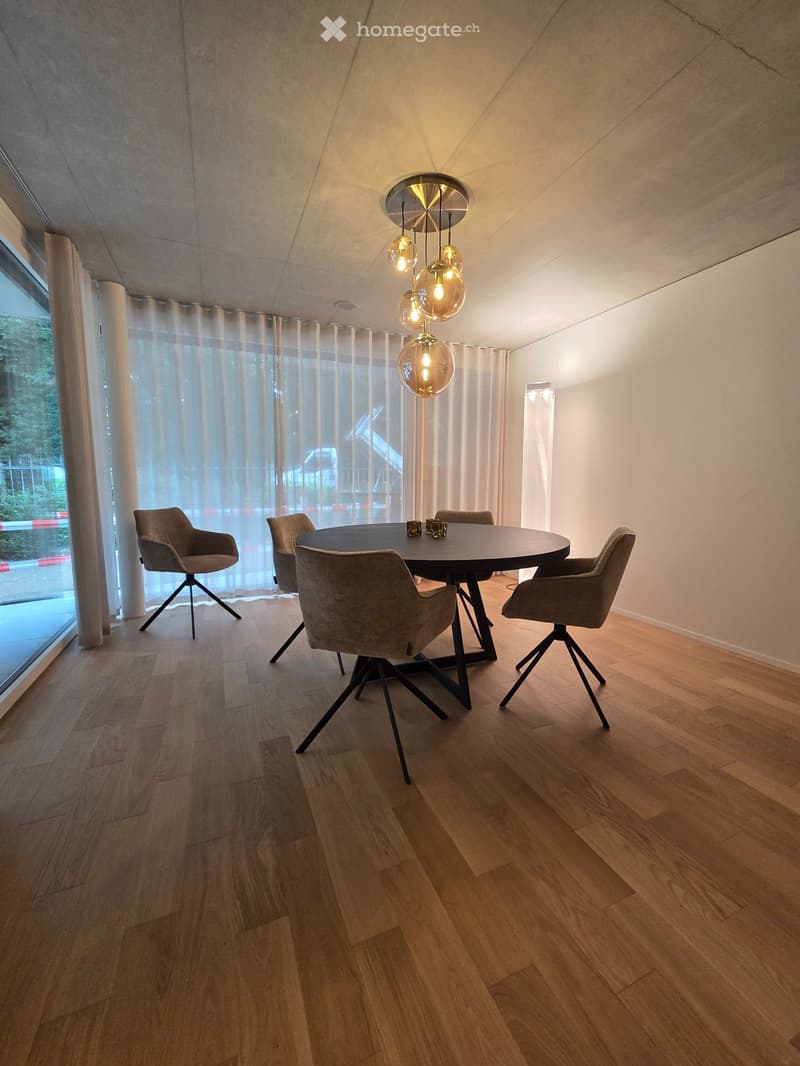 Modern dining room with a round wooden table, four grey chairs, pendant light fixture, and large window with sheer curtains.