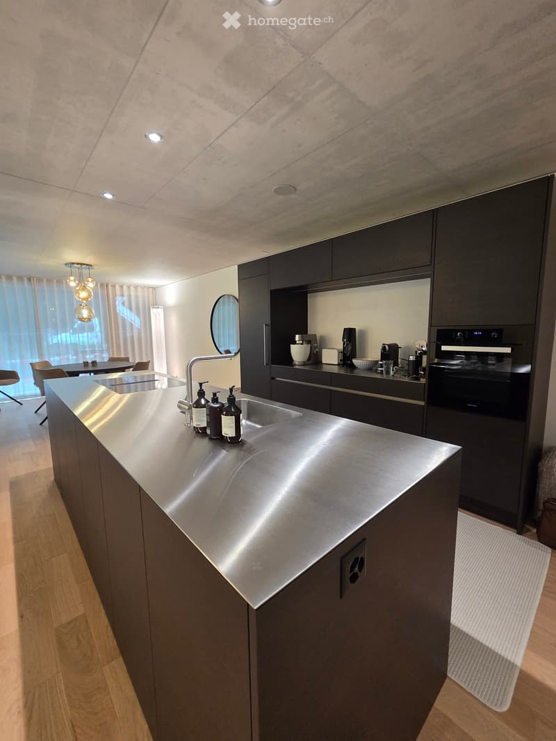 Modern kitchen with stainless steel island, dark cabinets, and built-in appliances. Features a sink, faucet, oven, coffee maker, blender, and a small seating area with chairs and a table.