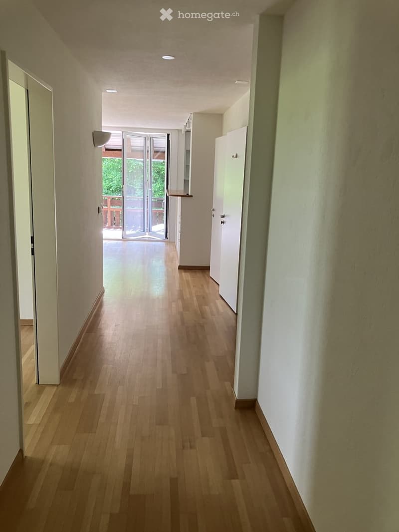 A long, well-lit hallway with wooden flooring, white walls, and several doors. A sliding glass door at the end leads to an outdoor area.