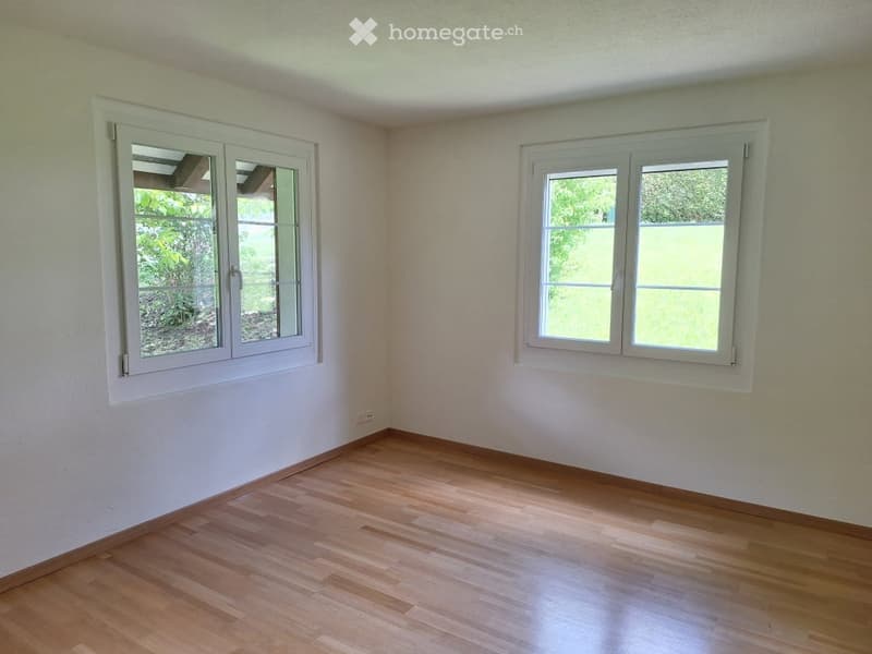 Empty room with light wooden flooring, white walls, and two large windows with white frames, offering a view of greenery outside.