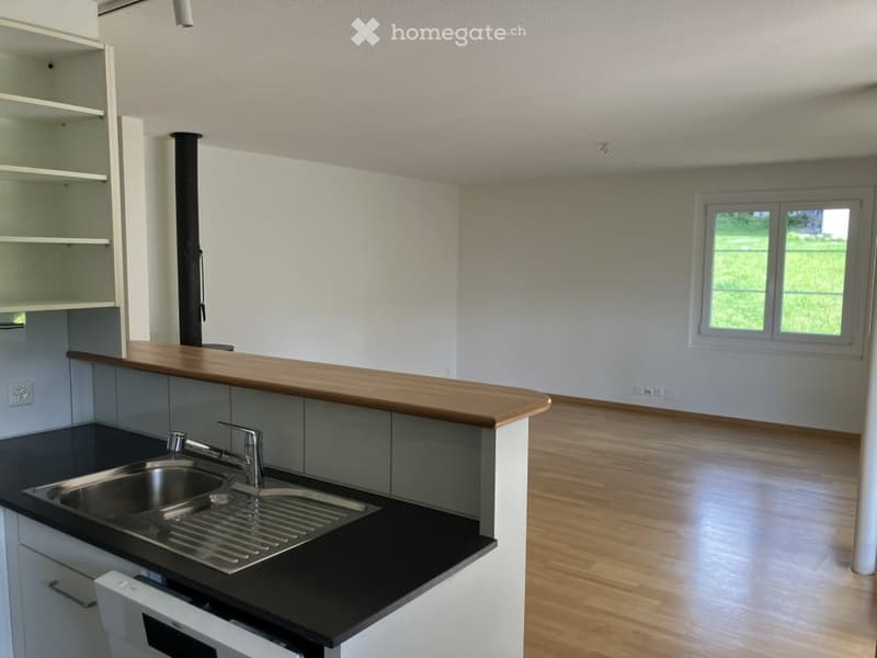 Empty kitchen with white cabinets, black countertops, a wooden island, and a stainless steel sink. The room features light-colored wooden flooring and large windows letting in natural light.