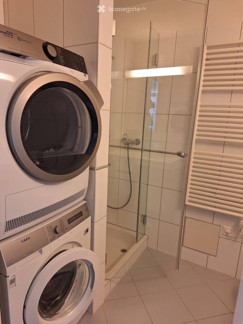 Modern bathroom with a glass-enclosed shower, a white towel rack, and stacked washing machine and dryer units.