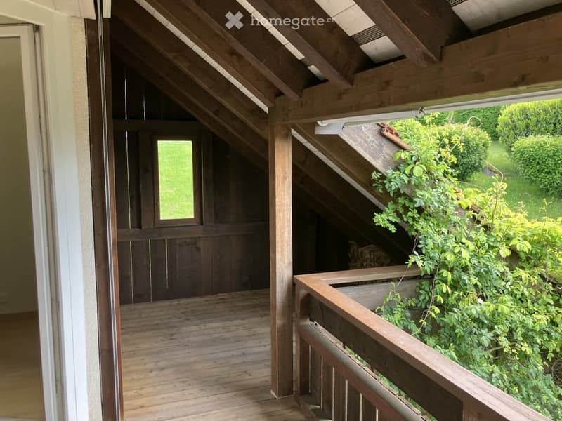 Wooden balcony with slanted wooden ceiling, railing, and view of greenery outside.