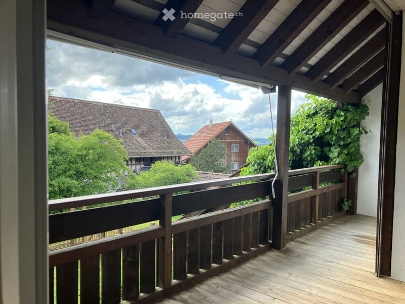 Wooden balcony with railing, overlooking a scenic countryside with houses and trees.