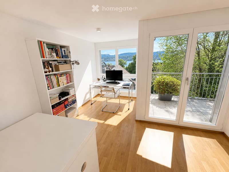 Bright, modern study with wooden flooring, large windows offering scenic views, a desk with a computer, a bookshelf filled with books, and a comfortable chair.