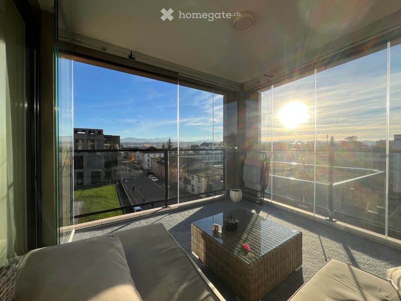 Modern balcony with large glass windows offering a scenic city view, equipped with a sofa, a wicker table, and a chair.