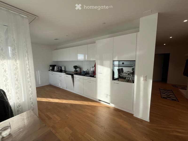 Modern kitchen with white cabinets, wooden flooring, and a small dining area visible through an open doorway.