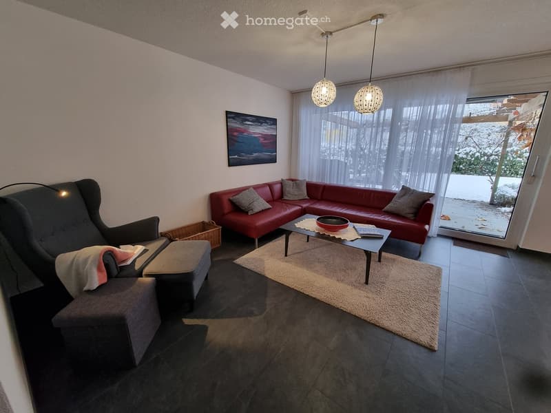 Modern living room with dark flooring, red sectional sofa, grey armchair, coffee table, and large windows with sheer curtains. Features a floor lamp, artwork, and a small basket near the chair.