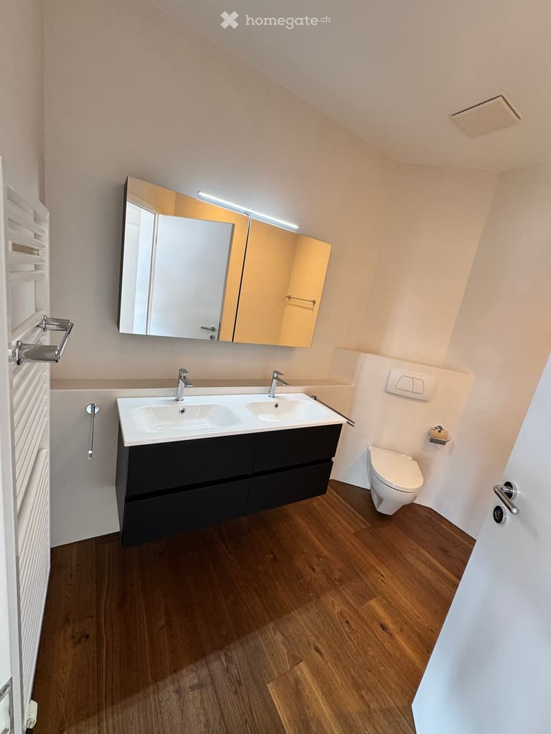 Bathroom with wooden floor, white walls, double sink, mirror, toilet, and towel rack