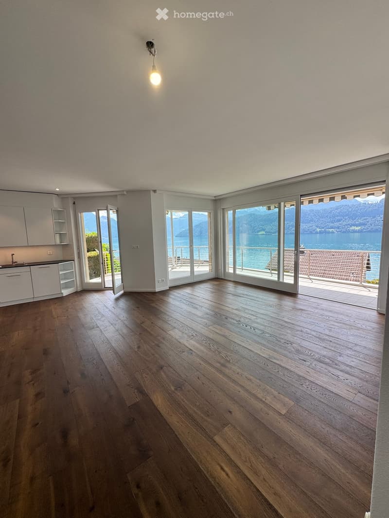 Empty room with wooden flooring, white walls, large windows, and a view of a lake and mountains.
