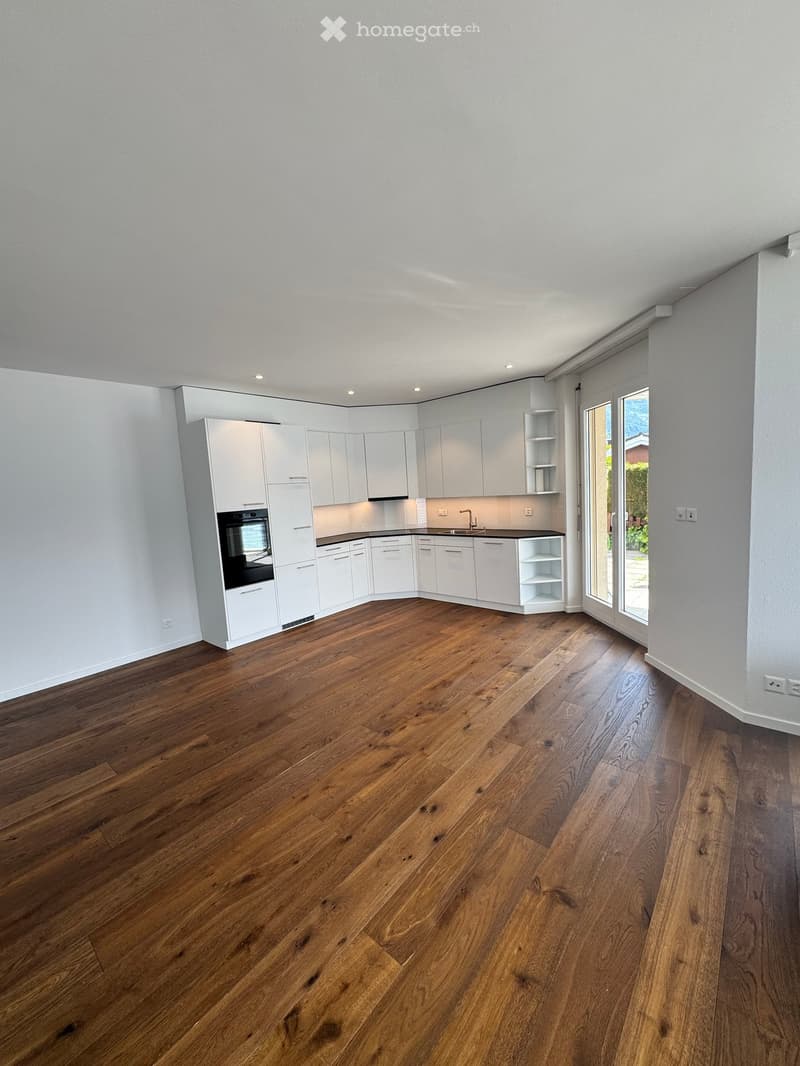 Empty kitchen with white cabinets, dark countertops, and wooden flooring. Features a built-in oven, sink, and large sliding glass door leading outside.