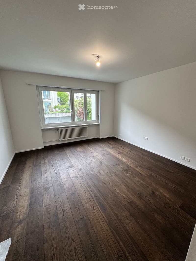 Empty room with wooden flooring, white walls, and a large window with a radiator below it.
