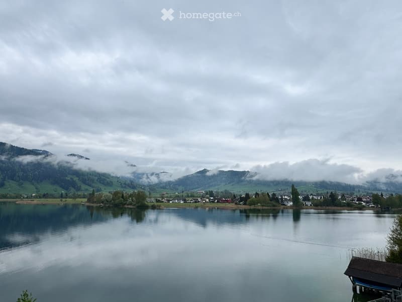 A serene lakeside view with calm water reflecting the surrounding landscape, featuring distant mountains partially shrouded in clouds and a small village along the shore.