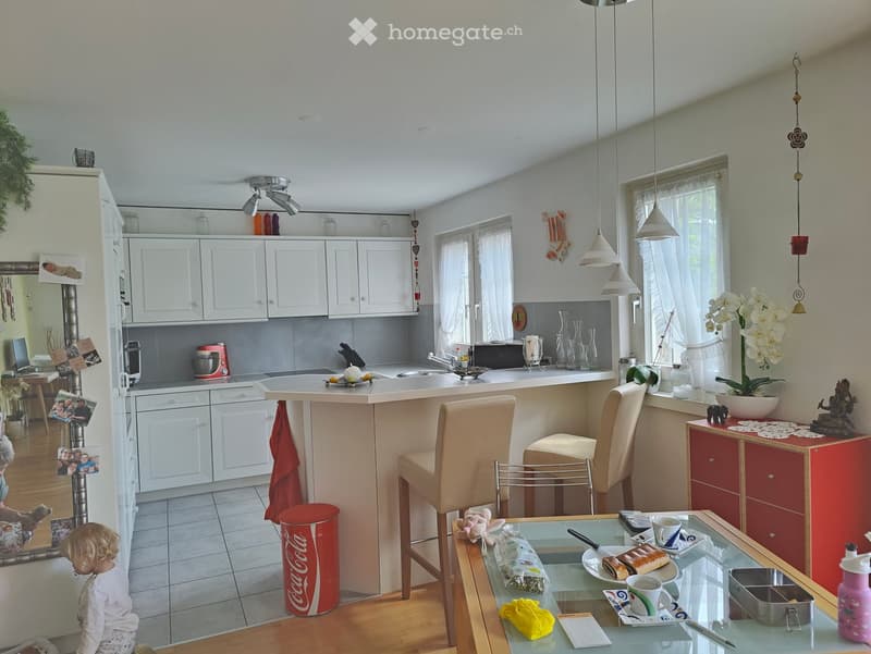 Bright, modern kitchen with white cabinets, a central island, and a dining area. Features include a refrigerator, sink, stools, and a glass table with various items on it.