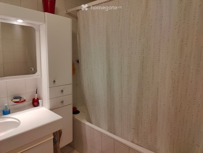 A small bathroom with a white sink, mirror, and cabinet. A shower curtain is drawn over a bathtub, and personal care items are placed on the sink counter.