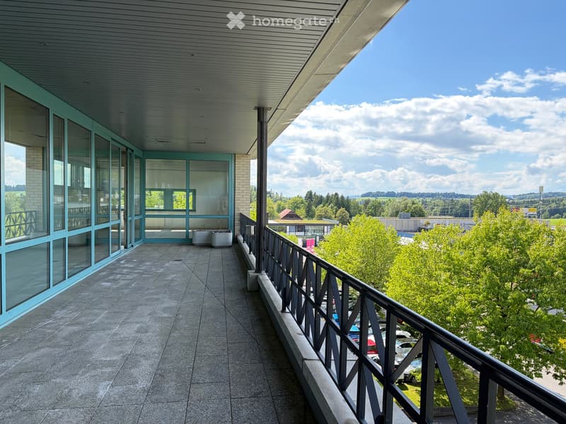 Balcony with modern glass walls, tiled floor, and metal railing overlooking a parking lot and greenery.