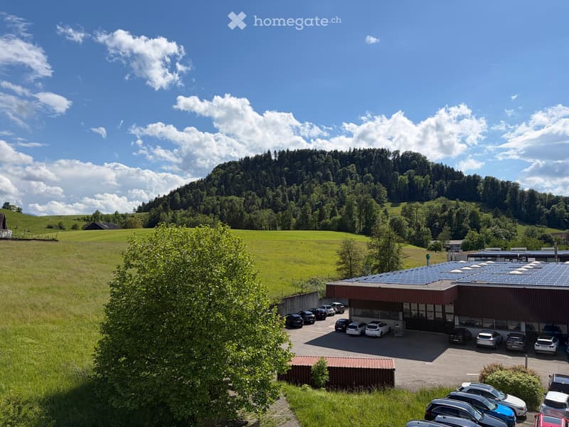 A modern building with a solar panel roof, surrounded by a parking lot with several cars, set in a lush green field with a forested hill in the background.