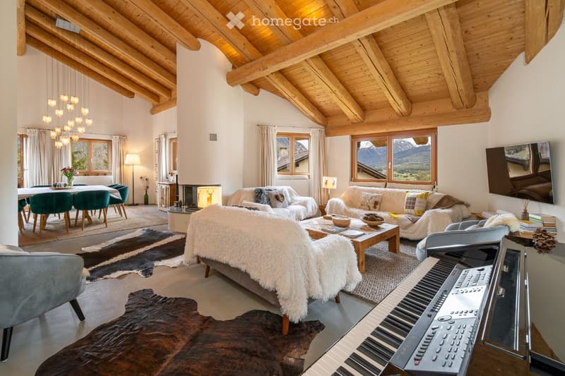 Spacious living room with exposed wooden beams, large windows offering a mountain view, a fireplace, a piano, a dining area with green chairs, and a cozy seating area with a fur throw on a couch.